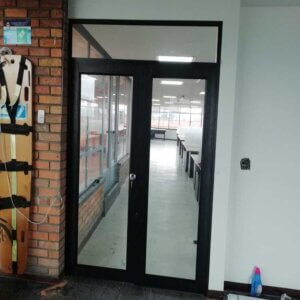 Puerta de emergencia de doble hoja con marcos negros de aluminio y paneles de vidrio templado, conectando un pasillo con pared de ladrillo y un interior de oficina moderna.